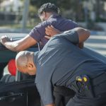 Agentes de segurança revistam uma pessoa ao lado de um carro durante uma abordagem policial.