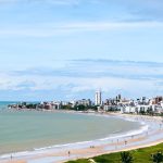 Vista aérea da orla de João Pessoa, com a faixa de areia, o mar e a área urbana ao fundo.