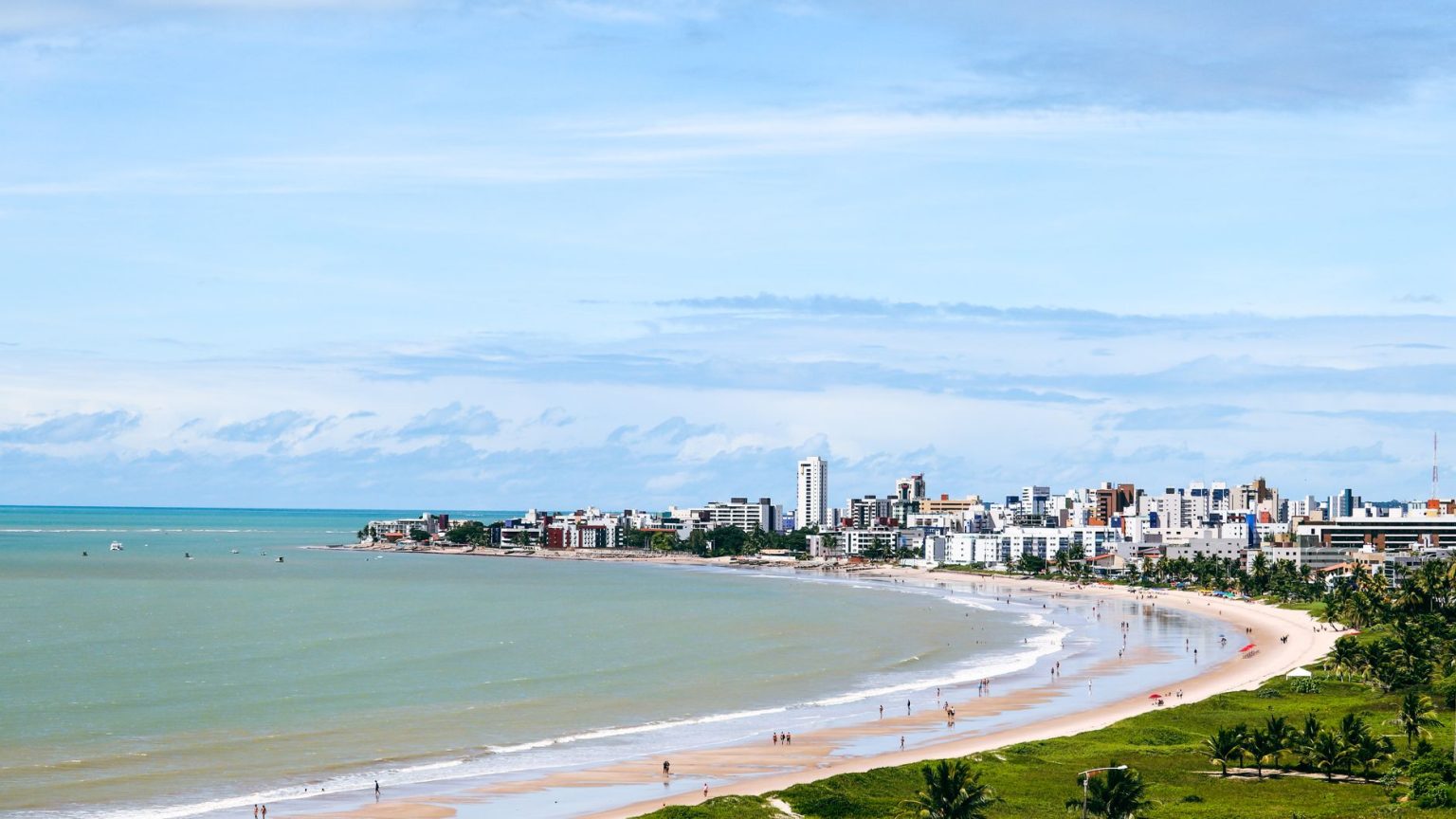 Vista aérea da orla de João Pessoa, com a faixa de areia, o mar e a área urbana ao fundo.