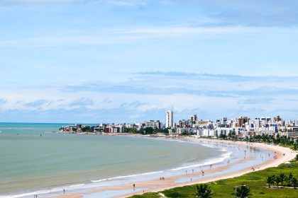 Vista aérea da orla de João Pessoa, com a faixa de areia, o mar e a área urbana ao fundo.
