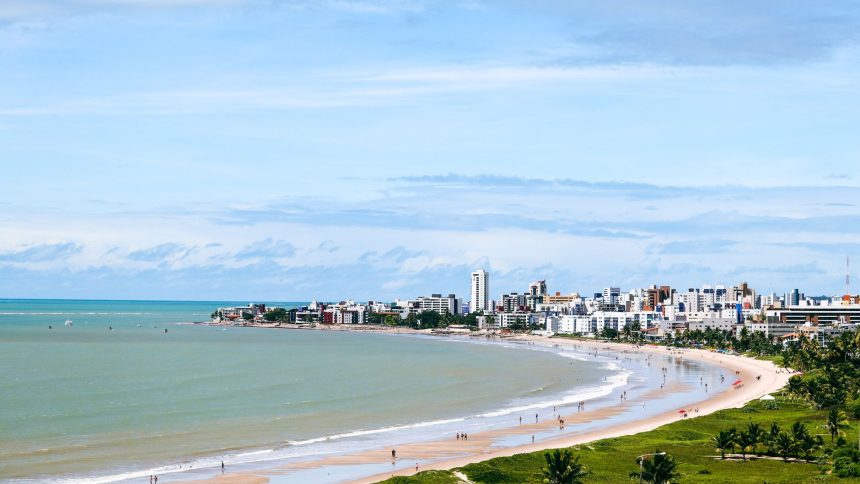 Vista aérea da orla de João Pessoa, com a faixa de areia, o mar e a área urbana ao fundo.