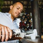 Homem serve uma bebida em um copo pequeno sobre um balcão de madeira, em um ambiente de bar.