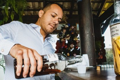 Homem serve uma bebida em um copo pequeno sobre um balcão de madeira, em um ambiente de bar.