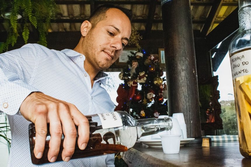 Homem serve uma bebida em um copo pequeno sobre um balcão de madeira, em um ambiente de bar.